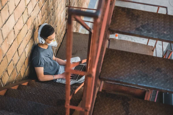 High angle view of asian teen in protective mask listening music with laptop on staircase, air pollution concept — Stock Photo