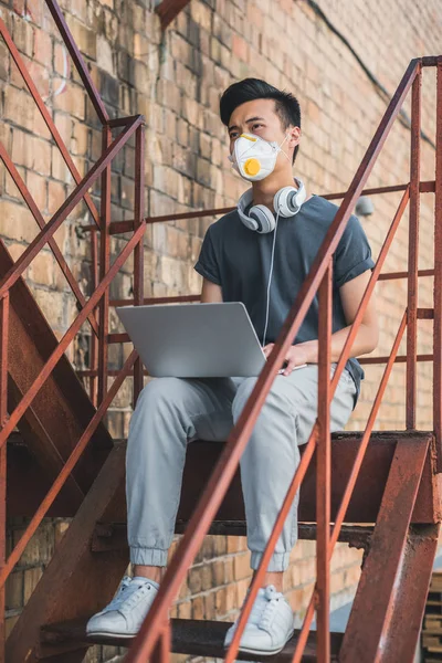 Asiatischer Teenager in Schutzmaske sitzt mit Laptop im Treppenhaus und schaut weg, Luftverschmutzungskonzept — Stockfoto