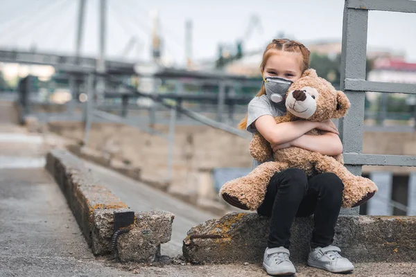 Criança em máscara protetora abraçando ursinho na rua, conceito de poluição do ar — Fotografia de Stock