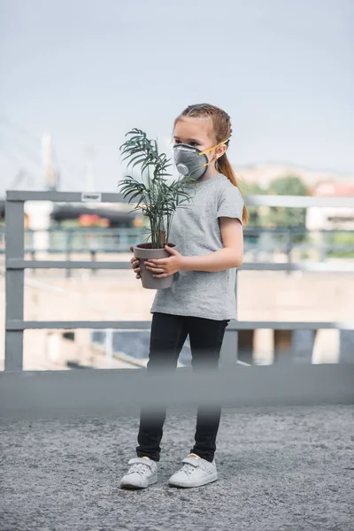 Criança em máscara protetora segurando planta envasada verde, conceito de poluição do ar — Fotografia de Stock