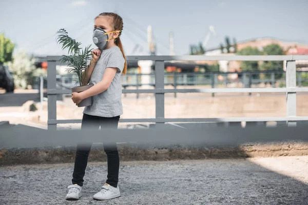 Child in protective mask carrying green potted plant, air pollution concept — Stock Photo