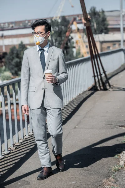 Asian businessman in protective mask walking on bridge with coffee to go and looking away, air pollution concept — Stock Photo