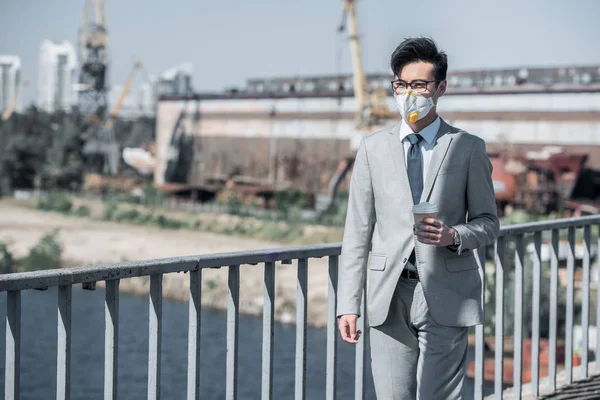 Asiatico uomo d'affari in maschera protettiva camminare su ponte con caffè in tazza di carta, inquinamento atmosferico concetto — Foto stock