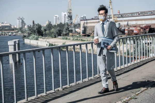 Asian businessman in protective mask walking on bridge with coffee in paper cup, air pollution concept — Stock Photo