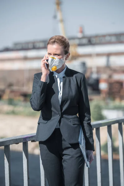 Предпринимательница в защитной маске разговаривает со смартфоном в промышленном районе, концепция загрязнения воздуха — стоковое фото