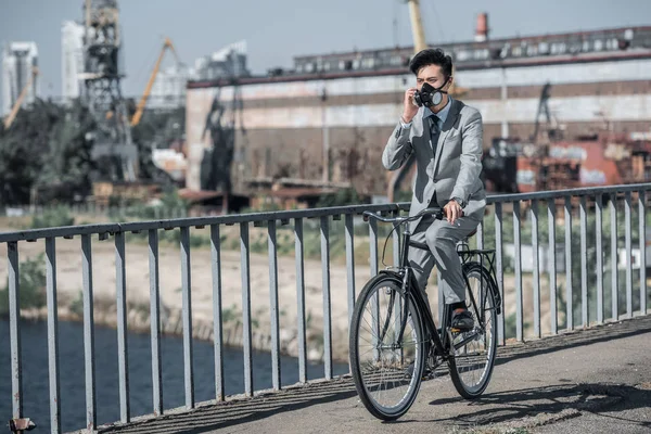 Азиатский бизнесмен в противогазе ездит на велосипеде по мосту и разговаривает по смартфону, концепция загрязнения воздуха — стоковое фото