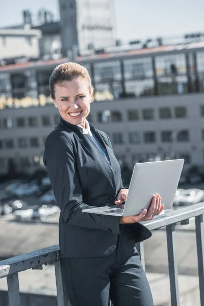 Smiling attractive businesswoman standing with laptop on bridge and looking at camera — Stock Photo