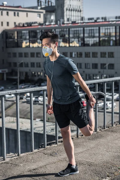 Sportive asian teenager stretching in protective mask on bridge, air pollution concept — Stock Photo