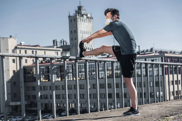 Азиатский подросток растягивает ноги в защитной маске на мосту, концепция загрязнения воздуха — стоковое фото