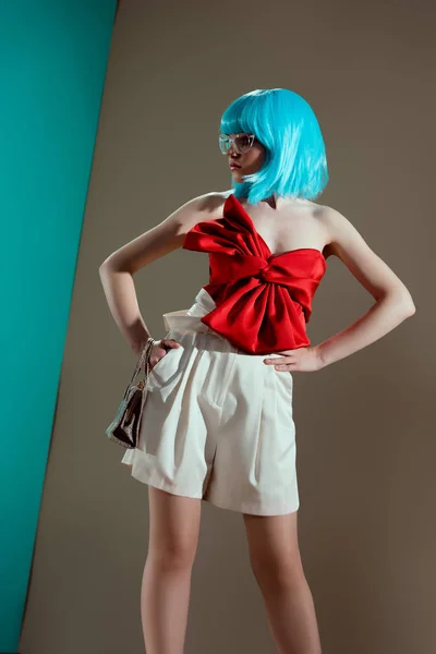 Belle fille à la mode en perruque bleue et arc rouge debout avec les mains sur la taille et regardant loin en studio — Photo de stock