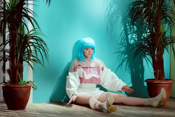 Schönes modisches Mädchen in blauer Perücke, das vorgibt, eine Puppe zu sein und auf dem Boden zwischen Topfpflanzen sitzt — Stockfoto