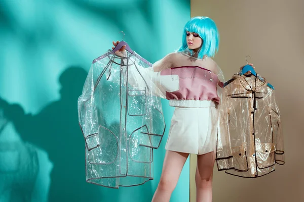 Hermosa chica de moda en peluca azul sosteniendo impermeables transparentes en el estudio - foto de stock