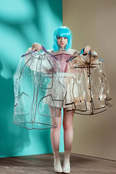 Beautiful girl in blue wig holding hangers with transparent raincoats and looking at camera — Stock Photo