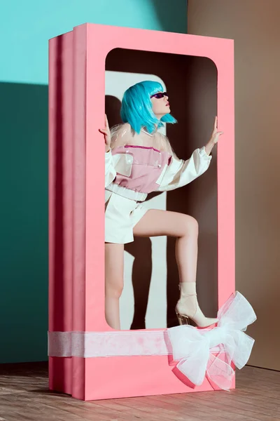 Chica hermosa de moda en peluca azul posando en caja rosa decorativa con lazo - foto de stock