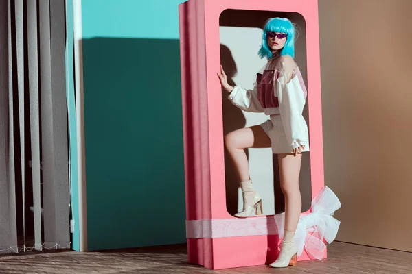 Fashionable young woman in blue wig posing in decorative pink box with bow — Stock Photo