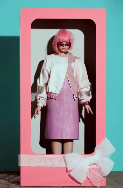 Schöne modische Mädchen in rosa Perücke steht in dekorativer Box mit Schleife und schaut in die Kamera — Stockfoto