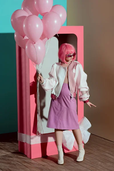 Модная девушка в розовом парике держа воздушные шары и глядя в сторону, стоя возле декоративной коробки с луком — стоковое фото
