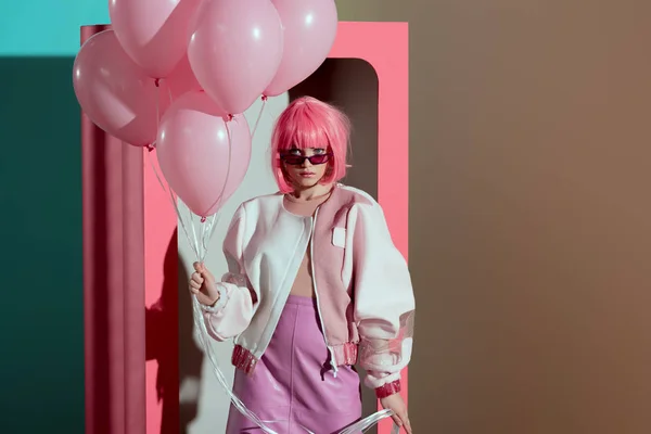 Chica con estilo en peluca rosa sosteniendo globos y mirando a la cámara mientras está de pie cerca de la caja decorativa - foto de stock