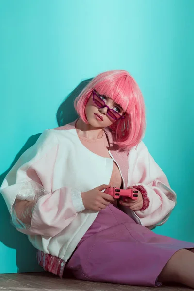 Chica bonita de moda en peluca rosa sentado en el suelo y jugando con joystick rosa - foto de stock