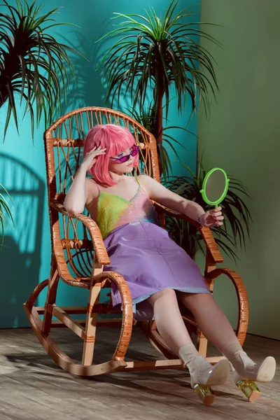 Модная молодая модель в розовом парике сидит в кресле-качалке и смотрит в зеркало — стоковое фото