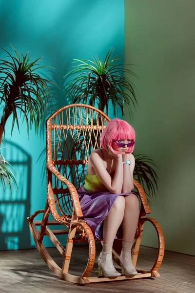 Belle fille ennuyée élégante en perruque rose assise dans un fauteuil à bascule et regardant loin — Photo de stock