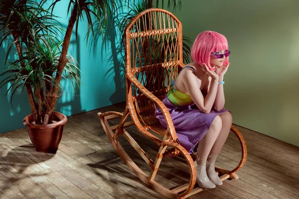 Hermosa chica con estilo en peluca rosa sentado en mecedora y mirando hacia otro lado - foto de stock