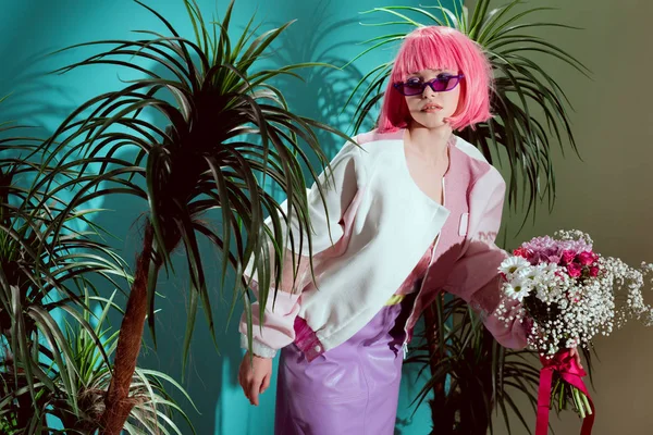 Hermosa mujer joven de moda en peluca rosa con ramo de flores y posando entre plantas de interior verdes - foto de stock