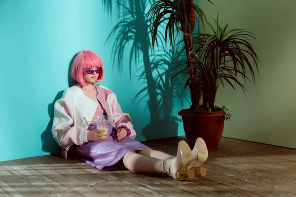 Attractive fashionable girl in pink wig holding disposable cup with smoothie and sitting on floor — Stock Photo