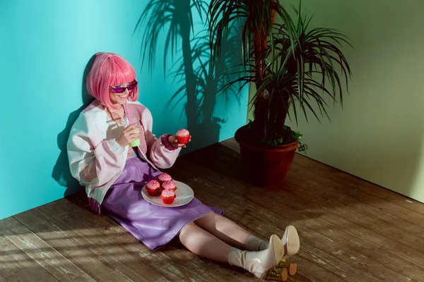 Sonriente chica en rosa peluca comer cupcakes y beber cóctel mientras está sentado en el suelo - foto de stock