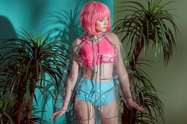 Hermosa chica elegante en peluca rosa y impermeable transparente mirando hacia otro lado entre las plantas verdes - foto de stock