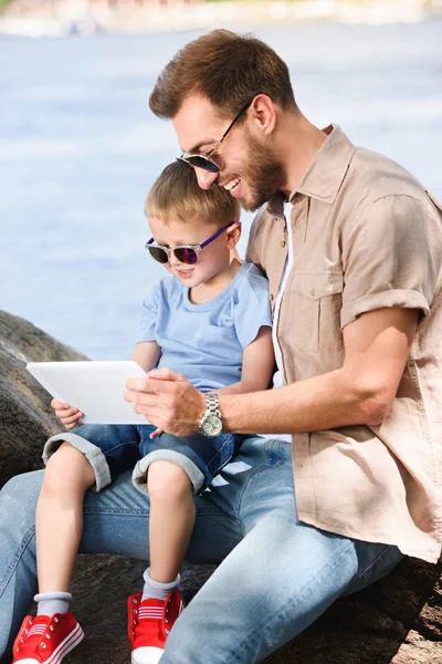 Padre e hijo usando la tableta cerca del río en el parque - foto de stock