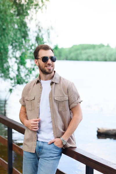 Sonriente hombre guapo apoyado en la barandilla cerca del lago en el parque - foto de stock