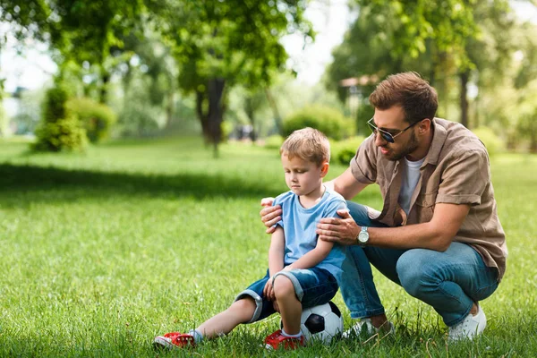 Отец обнимает и поддерживает грустного сына после игры в футбол в парке — стоковое фото