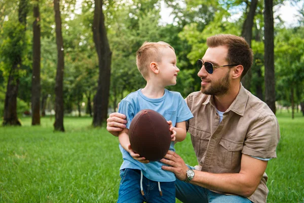 Glücklicher Vater und Sohn schauen einander an und halten American Football Ball im Park — Stockfoto