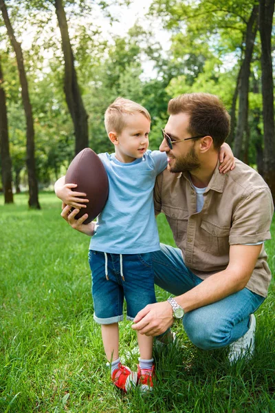 Padre e hijo abrazando y sosteniendo pelota de fútbol americano en el parque - foto de stock