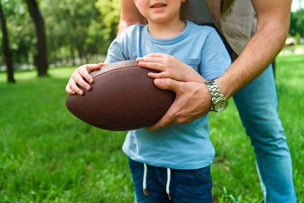 Imagen recortada de padre e hijo sosteniendo pelota de fútbol americano en el parque - foto de stock