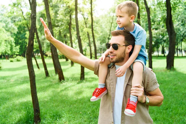 Батько махає рукою комусь і тримає сина на плечах в парку — стокове фото