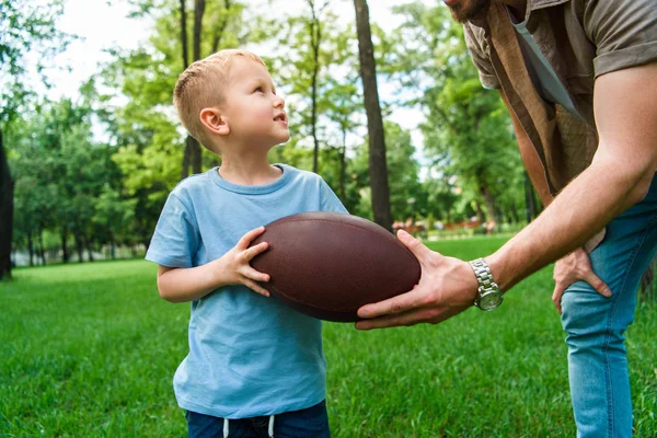 Imagen recortada de padre dando pelota de fútbol americano a hijo en el parque - foto de stock
