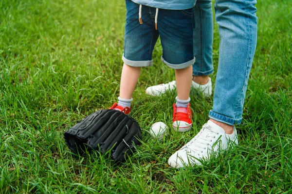 Image recadrée du père et du fils debout sur l'herbe près du ballon de baseball et du gant — Photo de stock