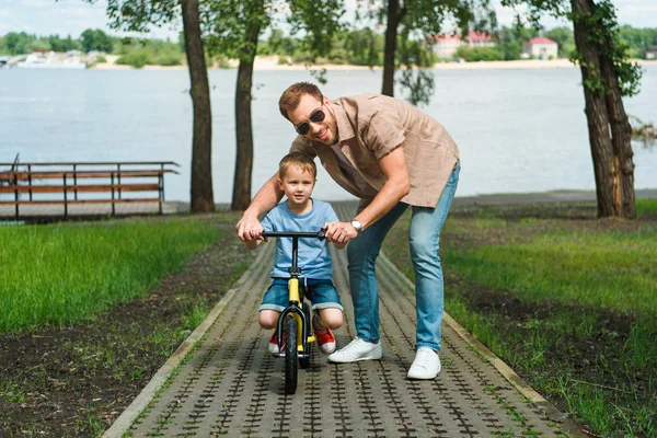 Father helping son riding small bike on road near river — Stock Photo