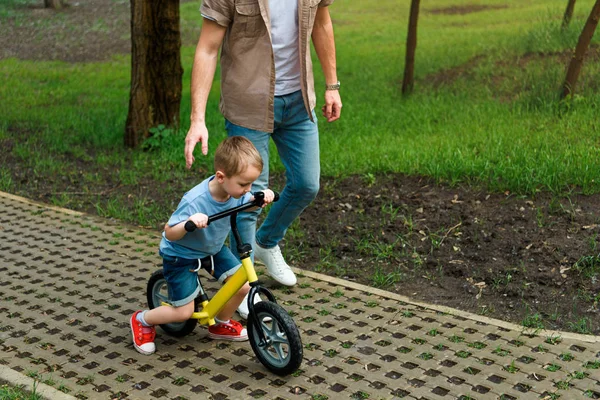 Immagine ritagliata del padre che sostiene il figlio mentre va in bicicletta al parco — Foto stock