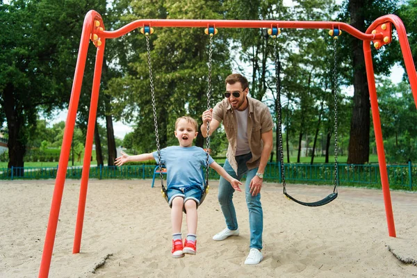 Отец и сын веселятся на качелях на детской площадке в парке — стоковое фото