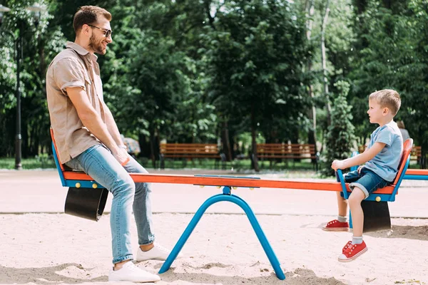 Vista lateral de pai e filho se divertindo no balanço no parque infantil no parque — Fotografia de Stock