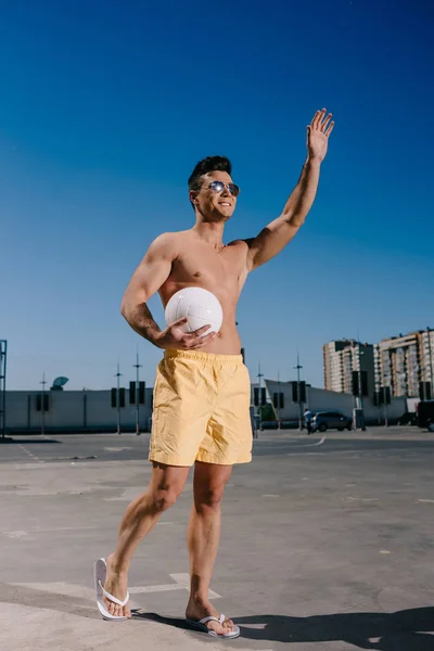 Hombre feliz sin camisa sosteniendo la pelota de voleibol y saludando la mano en el estacionamiento - foto de stock