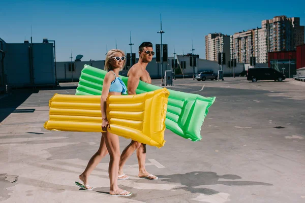 Jeune couple en vêtements de plage avec lits gonflables marchant sur le parking — Photo de stock