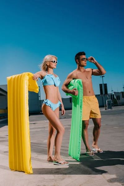 Jeune couple en vêtements de plage avec lits gonflables debout sur le parking — Photo de stock