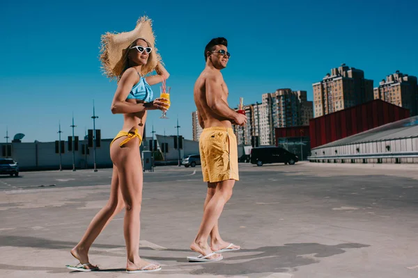 Молодая счастливая пара в купальниках с освежающими коктейлями, стоящими на парковке — стоковое фото