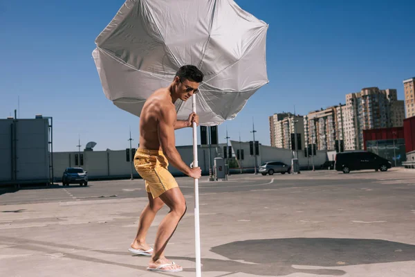 Masculine young shirtless man putting beach umbrella in asphalt on parking — Stock Photo