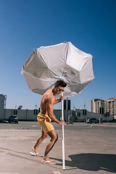Atlético joven sin camisa hombre poner paraguas de playa en asfalto en el aparcamiento - foto de stock