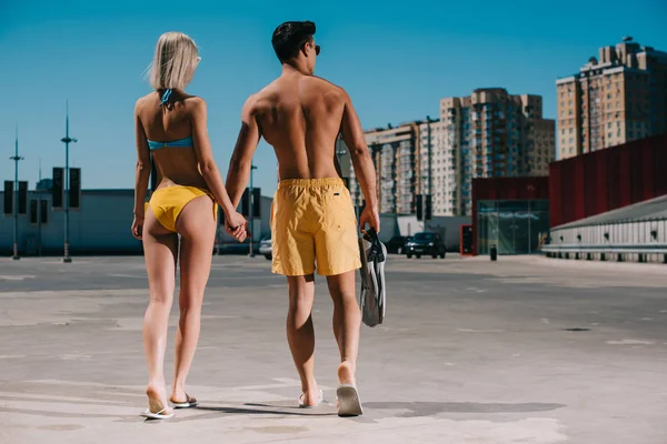 Rear view of young couple in bikini and swimming shorts with flippers walking on parking — Stock Photo